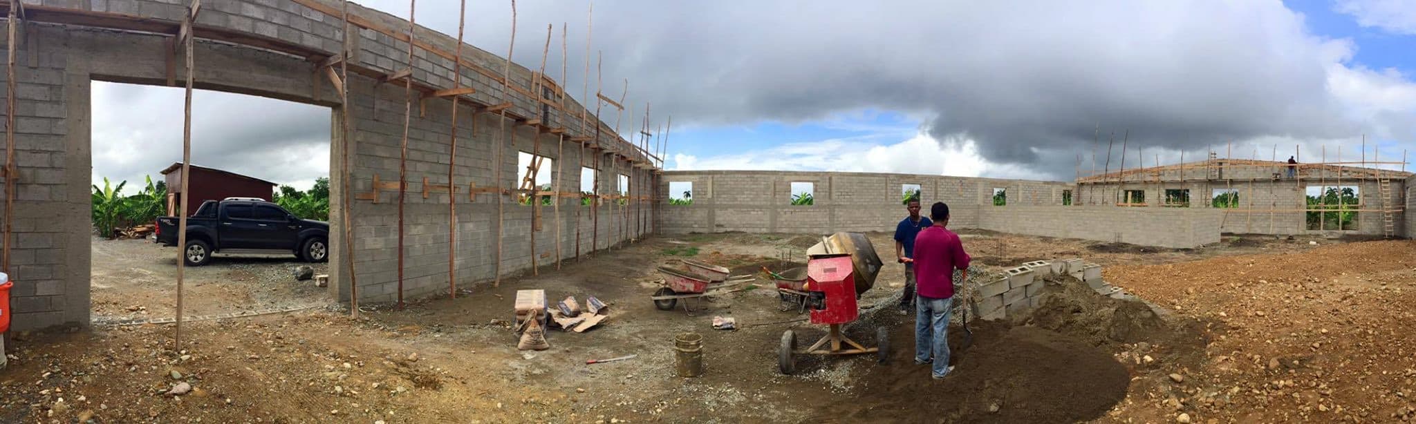 Wide view of masonry walls, timber scaffolding, and builders across the M2535 Vocational Village construction site