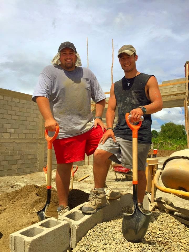 Two volunteer builders standing with shovels beside concrete blocks and a mixer at the M2535 construction site