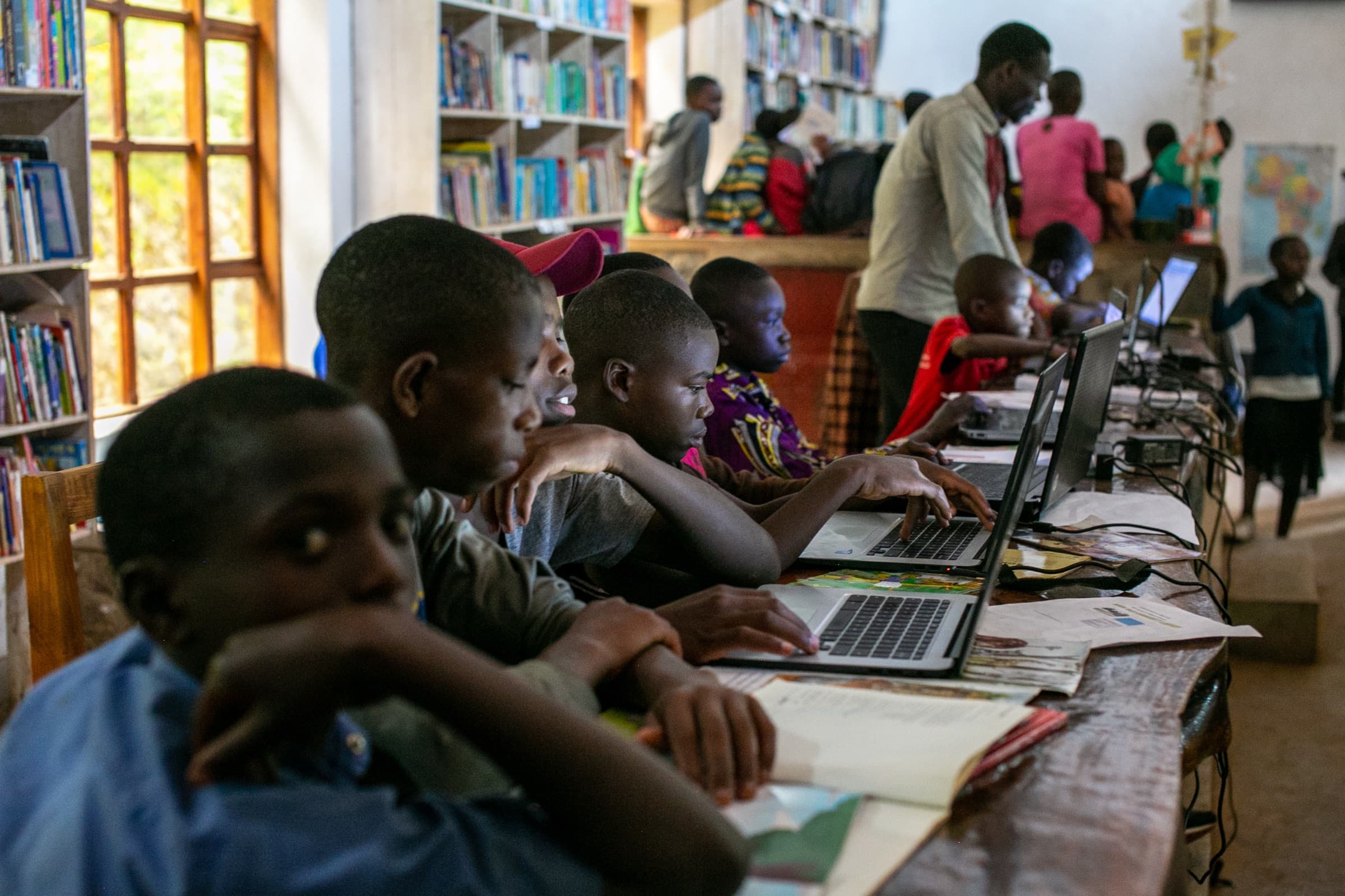 Children working together on laptops between the bookshelves of the Sunzu Village library